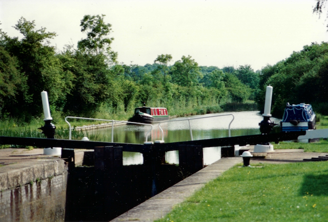 A tranquil canal scene featuring a narrowboat positioned in a lock, surrounded by lush green foliage and trees.