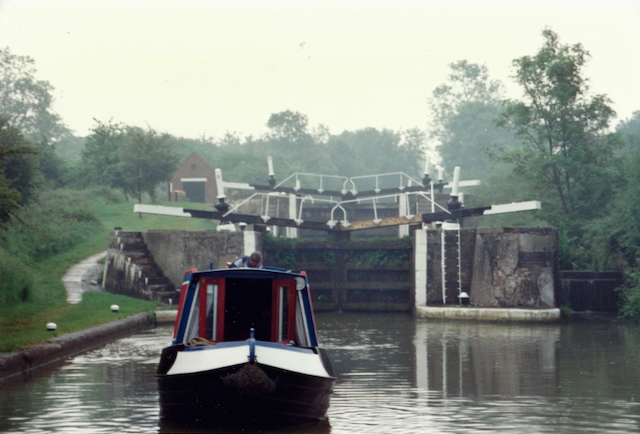 A narrowboat approaching a canal lock surrounded by greenery on a misty day.